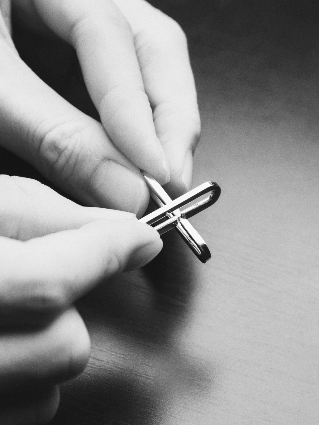A close-up black and white photo of hands holding a small metallic cross-shaped accessory over a dark wooden surface.