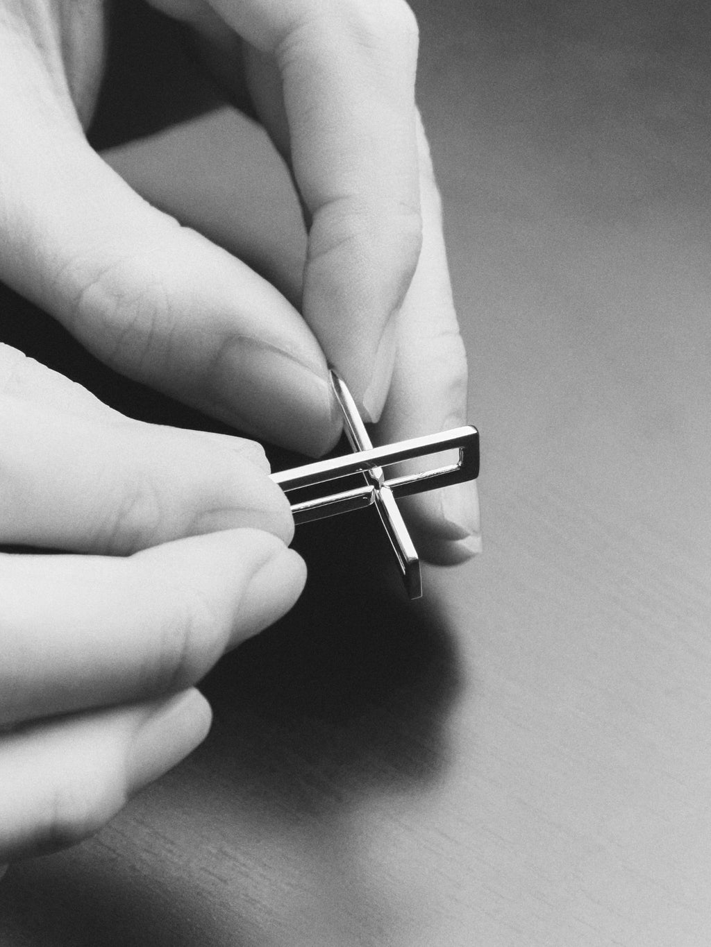 A close-up black and white photo of a person's hands holding a small metallic cross-shaped accessory, likely a cufflink or pendant, over a dark surface.